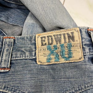 May include: Close-up of blue denim jeans with an EDWIN label. The label is rectangular with the brand name in a dark font and turquoise text. Orange stitching is visible on the jeans.