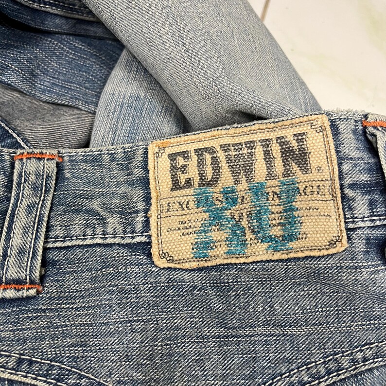 May include: Close-up of blue denim jeans with an EDWIN label. The label is rectangular with the brand name in a dark font and turquoise text. Orange stitching is visible on the jeans.