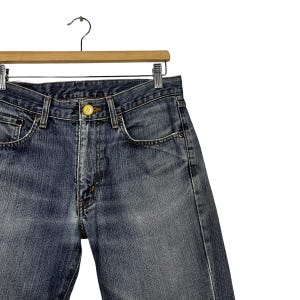 May include: Close-up of blue denim jeans hanging on a wooden hanger. The jeans feature a button closure and a classic five-pocket design. The denim has a faded wash, with visible wear and tear.