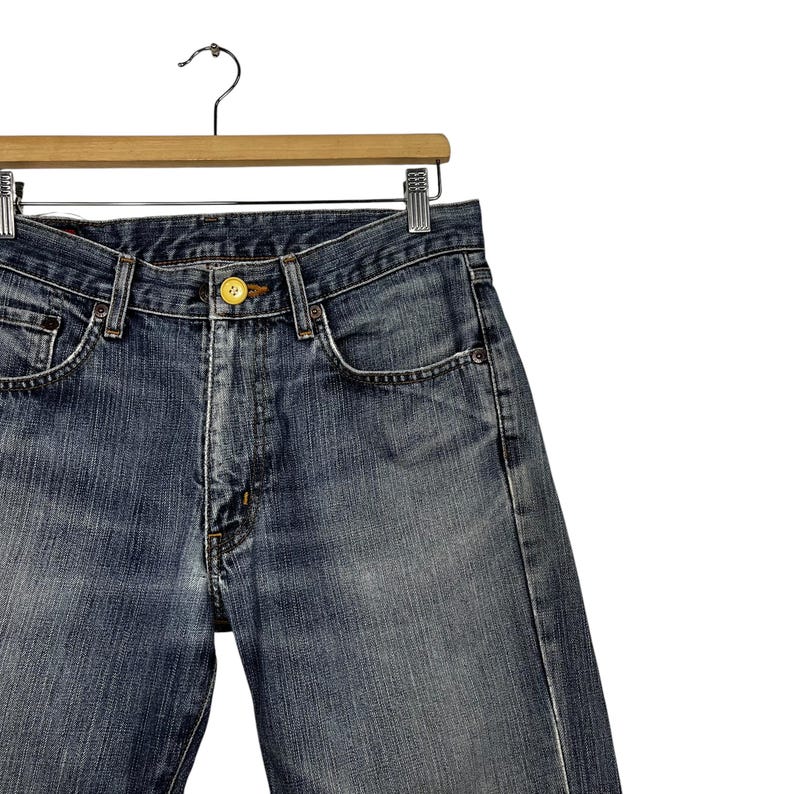 May include: Close-up of blue denim jeans hanging on a wooden hanger. The jeans feature a button closure and a classic five-pocket design. The denim has a faded wash, with visible wear and tear.