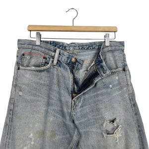 May include: Light blue denim jeans with a distressed look, featuring a button closure and zipper. The jeans have a worn appearance with visible distressing and a few small holes. They are hanging on a wooden hanger.