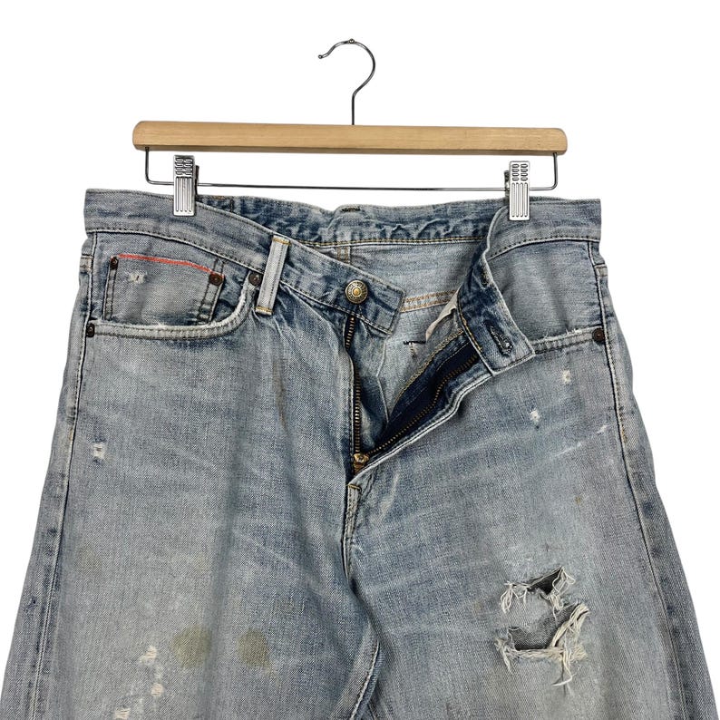 May include: Light blue denim jeans with a distressed look, featuring a button closure and zipper. The jeans have a worn appearance with visible distressing and a few small holes. They are hanging on a wooden hanger.