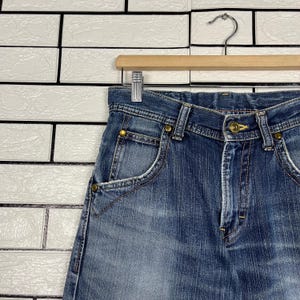 May include: Close-up of blue denim jeans with a classic five-pocket design. The jeans are hanging on a wooden hanger against a white brick wall. The jeans have a button and zipper closure, and brass-colored rivets.