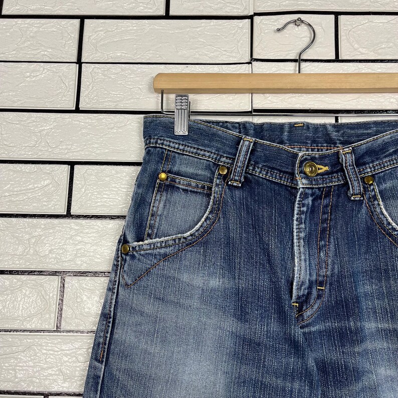 May include: Close-up of blue denim jeans with a classic five-pocket design. The jeans are hanging on a wooden hanger against a white brick wall. The jeans have a button and zipper closure, and brass-colored rivets.
