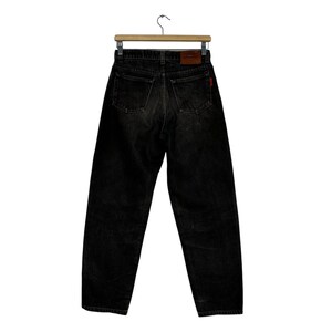 Puede incluir: Vaqueros de mezclilla negros colgados de una percha de madera. Los vaqueros tienen un dise&ntilde;o cl&aacute;sico de cinco bolsillos, una etiqueta roja y un parche de cuero marr&oacute;n con un logotipo. Los vaqueros son de lavado oscuro y parecen vintage.