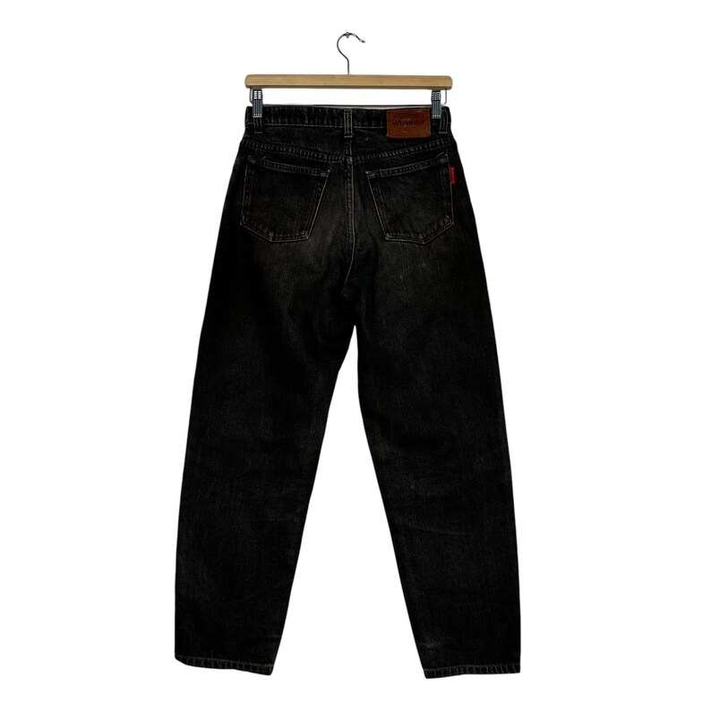 Puede incluir: Vaqueros de mezclilla negros colgados de una percha de madera. Los vaqueros tienen un dise&ntilde;o cl&aacute;sico de cinco bolsillos, una etiqueta roja y un parche de cuero marr&oacute;n con un logotipo. Los vaqueros son de lavado oscuro y parecen vintage.