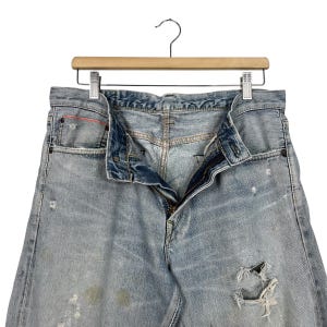 May include: Light blue denim jeans with distressed details, hanging on a wooden hanger. The jeans have a button and zipper closure, with visible wear and tear, including a ripped section on the leg.