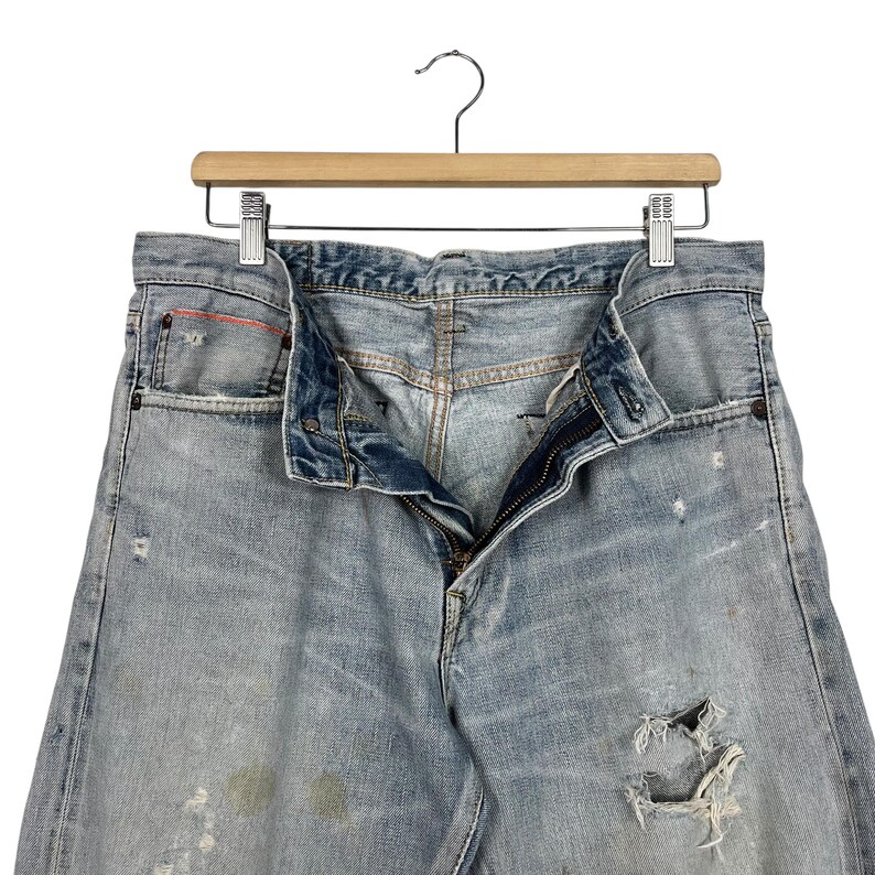 May include: Light blue denim jeans with distressed details, hanging on a wooden hanger. The jeans have a button and zipper closure, with visible wear and tear, including a ripped section on the leg.
