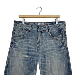 May include: Light blue denim jeans with a distressed wash, featuring classic five-pocket styling and red stitching accents. The jeans are hanging on a wooden hanger against a white background.