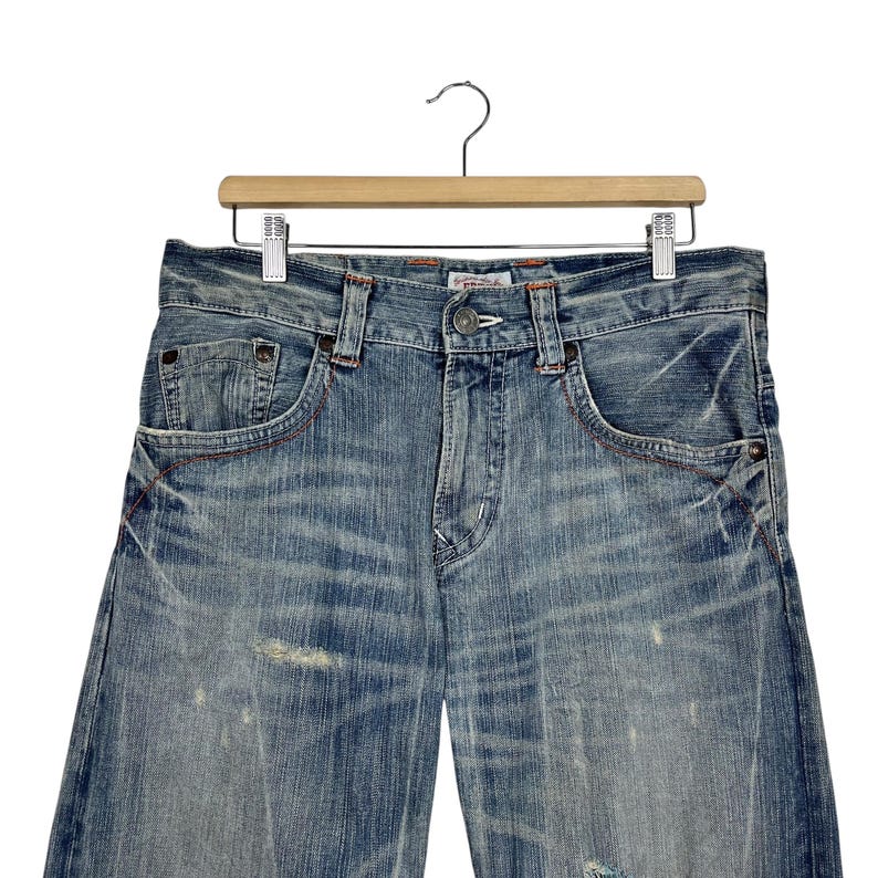 May include: Light blue denim jeans with a distressed wash, featuring classic five-pocket styling and red stitching accents. The jeans are hanging on a wooden hanger against a white background.