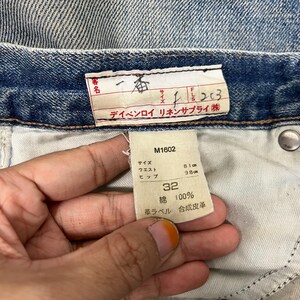 K&ouml;nnte beinhalten: Nahaufnahme der Innenseite einer blauen Jeans. Ein wei&szlig;es Etikett zeigt die Gr&ouml;&szlig;e 32, mit Taillen- und H&uuml;ftma&szlig;en in cm. Das Etikett gibt auch an, dass die Jeans aus 100 % Baumwolle besteht.
