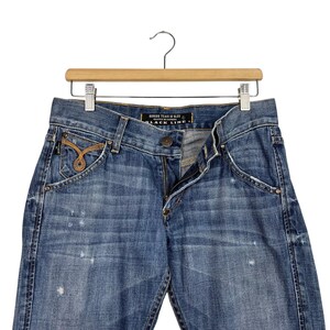 K&ouml;nnte beinhalten: Blaue Jeans aus Denim mit einem Used-Look, h&auml;ngen an einem Holzkleiderb&uuml;gel. Die Jeans haben einen Knopf- und Rei&szlig;verschluss, Vordertaschen und dekorative N&auml;hte. Das Etikett lautet "BLACK LINE".