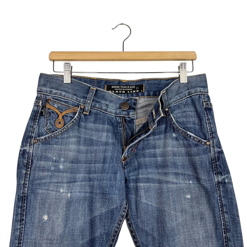 K&ouml;nnte beinhalten: Blaue Jeans aus Denim mit einem Used-Look, h&auml;ngen an einem Holzkleiderb&uuml;gel. Die Jeans haben einen Knopf- und Rei&szlig;verschluss, Vordertaschen und dekorative N&auml;hte. Das Etikett lautet "BLACK LINE".