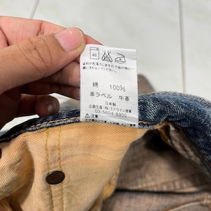 May include: A close-up of a brown leather label with white text that reads "100% Cotton, Leather Label, Cowhide, Made in Japan, (Stock) Edwin Trading Co., 03-5604-8900, Caution: Sweat or rain, and friction."