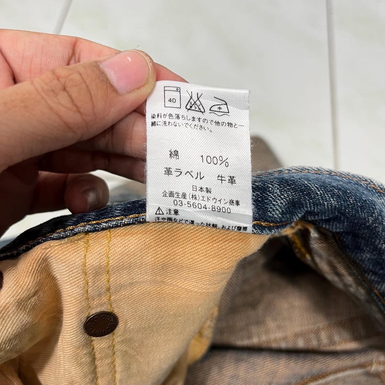 May include: A close-up of a brown leather label with white text that reads "100% Cotton, Leather Label, Cowhide, Made in Japan, (Stock) Edwin Trading Co., 03-5604-8900, Caution: Sweat or rain, and friction."