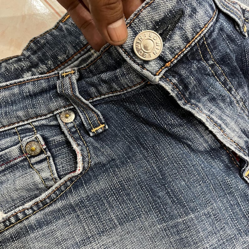 Op de afbeelding: Close-up van een blauwe spijkerbroek met zichtbare stiksels en metalen knoopdetails. De jeans heeft een klassiek ontwerp met vijf zakken, met een knoopsluiting en een ritssluiting. De denimstof vertoont tekenen van slijtage.