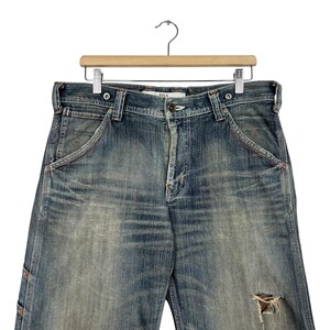 May include: Blue denim jeans with a distressed appearance, hanging on a wooden hanger. The jeans show signs of wear and tear, including a distressed area on the right leg. The stitching is in a contrasting orange colour.