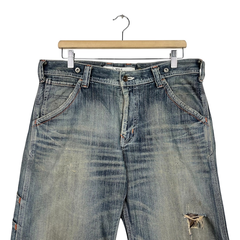 May include: Blue denim jeans with a distressed appearance, hanging on a wooden hanger. The jeans show signs of wear and tear, including a distressed area on the right leg. The stitching is in a contrasting orange colour.