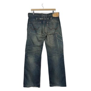 May include: Faded blue denim jeans with a carpenter pocket and a leather patch. The jeans are hanging on a wooden hanger. The back pockets have a red stitch design.