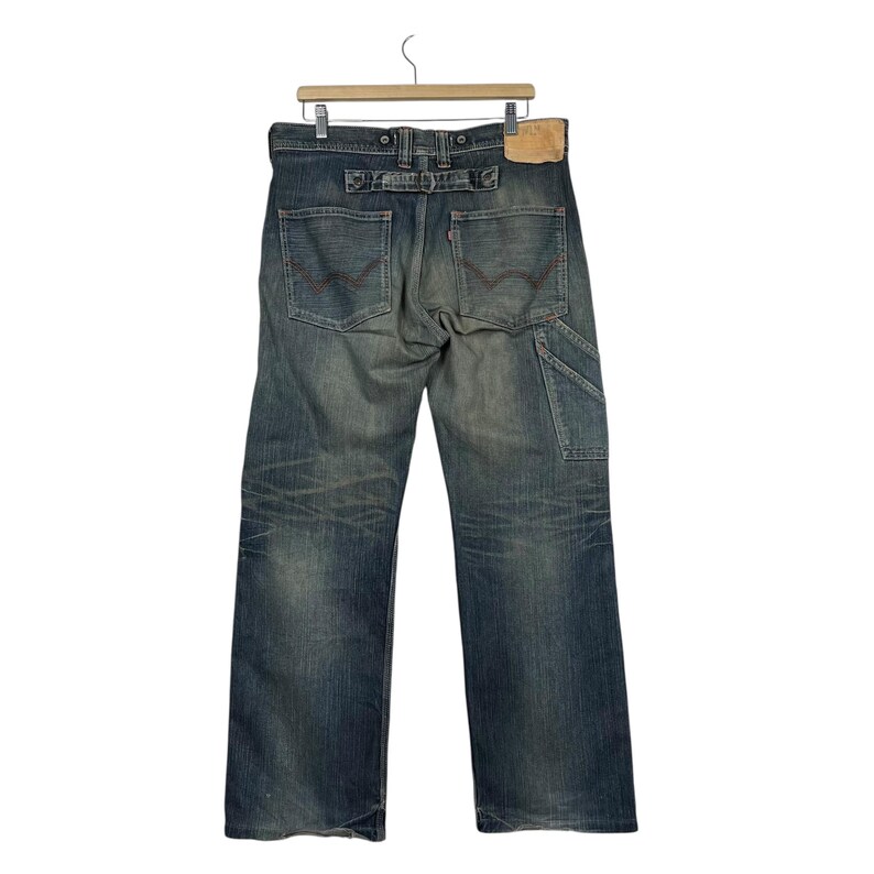 May include: Faded blue denim jeans with a carpenter pocket and a leather patch. The jeans are hanging on a wooden hanger. The back pockets have a red stitch design.