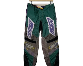 Op de afbeelding: Groene, paarse en grijze motorbroek met het MSR-logo. De broek heeft zwarte accenten en hangt aan een houten hanger. Ontworpen voor offroad motorrijden.