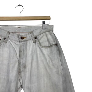 May include: Light gray denim jeans hanging on a wooden hanger. The jeans have a classic five-pocket design with a button and zipper closure. The denim has a faded, worn appearance.