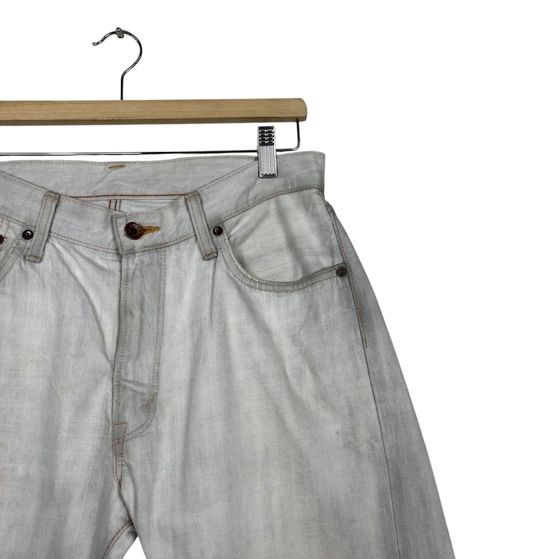 May include: Light gray denim jeans hanging on a wooden hanger. The jeans have a classic five-pocket design with a button and zipper closure. The denim has a faded, worn appearance.