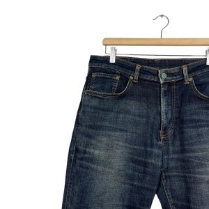 Puede incluir: Vaqueros de mezclilla de lavado oscuro colgados de una percha de madera. Los vaqueros tienen un dise&ntilde;o cl&aacute;sico de cinco bolsillos con cierre de bot&oacute;n y cremallera. La mezclilla tiene un aspecto descolorido y desgastado.