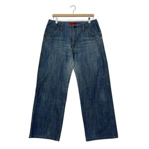 May include: Blue denim jeans with a wide leg design, hanging from a wooden hanger. The jeans have a classic five-pocket design, a button and zipper closure, and a red tag on the waistband. The jeans are a medium wash with subtle fading.