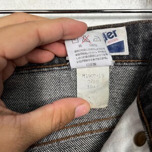 Puede incluir: Primer plano de unos vaqueros negros. Los vaqueros tienen una etiqueta blanca con la marca "Wrangler Goods" y texto japon&eacute;s. Una etiqueta m&aacute;s peque&ntilde;a muestra el texto "M1907-19" y otros n&uacute;meros. Los vaqueros son 100% algod&oacute;n.