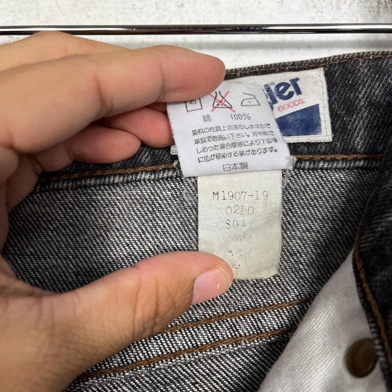 Puede incluir: Primer plano de unos vaqueros negros. Los vaqueros tienen una etiqueta blanca con la marca "Wrangler Goods" y texto japon&eacute;s. Una etiqueta m&aacute;s peque&ntilde;a muestra el texto "M1907-19" y otros n&uacute;meros. Los vaqueros son 100% algod&oacute;n.