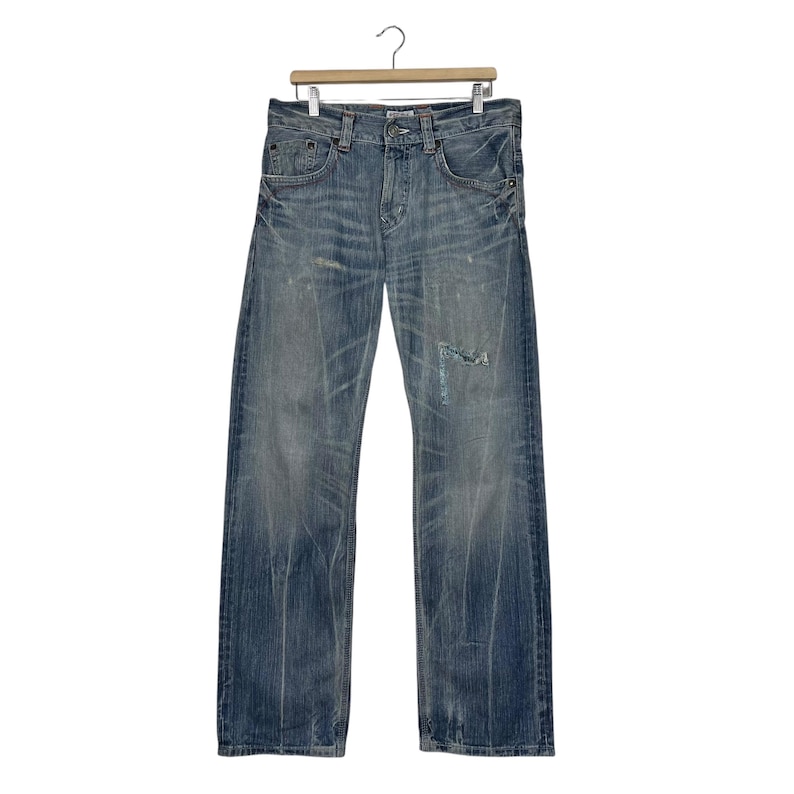 May include: Light blue denim jeans with a distressed look, featuring fading and a ripped knee. The jeans are hanging on a wooden hanger against a white background. The jeans have a classic five-pocket design.