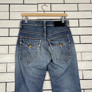 May include: Blue denim jeans with a classic five-pocket design, featuring a button and zipper closure. The back pockets have a distinctive embroidered "W" logo. The jeans are hanging on a wooden hanger.