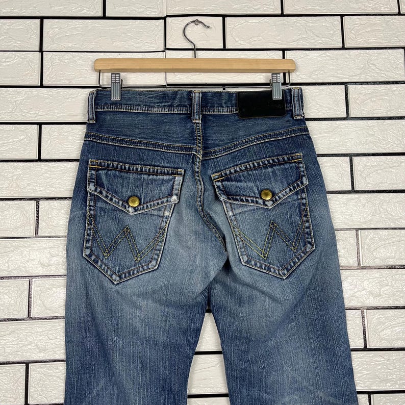 May include: Blue denim jeans with a classic five-pocket design, featuring a button and zipper closure. The back pockets have a distinctive embroidered "W" logo. The jeans are hanging on a wooden hanger.
