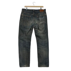 May include: Faded blue denim jeans hanging on a wooden hanger. The jeans have a classic five-pocket design, with a worn appearance and subtle fading throughout. A brown leather patch is visible on the back.