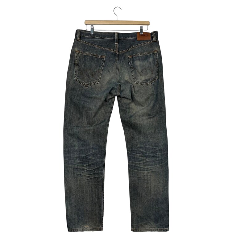 May include: Faded blue denim jeans hanging on a wooden hanger. The jeans have a classic five-pocket design, with a worn appearance and subtle fading throughout. A brown leather patch is visible on the back.