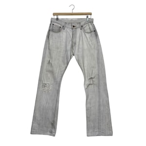 May include: Light gray denim jeans with distressed details, including rips at the knees and thighs. The jeans are hanging on a wooden hanger against a white background. The jeans have a classic five-pocket design.