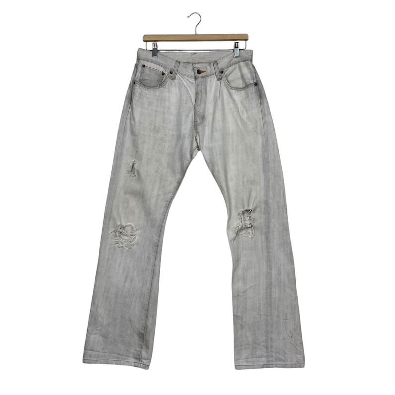 May include: Light gray denim jeans with distressed details, including rips at the knees and thighs. The jeans are hanging on a wooden hanger against a white background. The jeans have a classic five-pocket design.