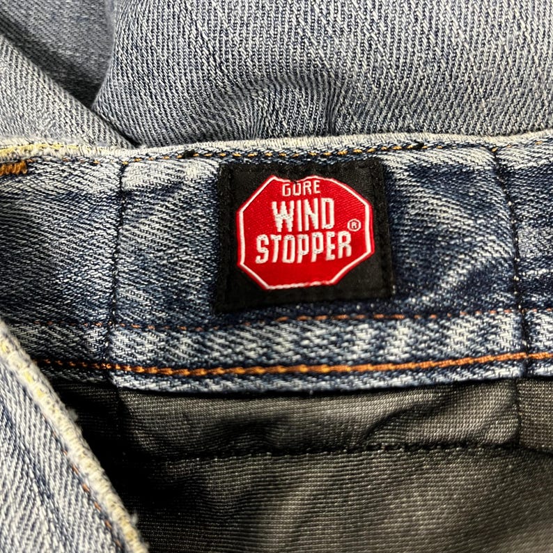 K&ouml;nnte beinhalten: Nahaufnahme von blauem Denim-Stoff mit einem rot-wei&szlig;en "GORE WIND STOPPER"-Etikett. Das Etikett hat die Form eines Stoppschilds, und der Denim hat sichtbare N&auml;hte und ein strukturiertes Aussehen.