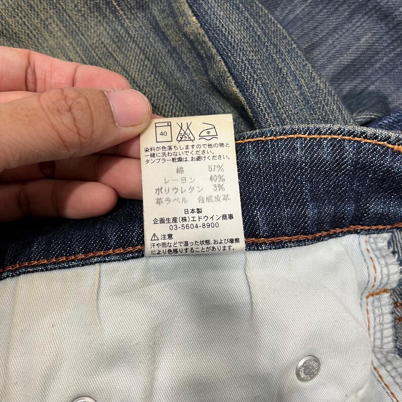K&ouml;nnte beinhalten: Nahaufnahme eines Jeansetiketts mit japanischem Text und Pflegesymbolen. Das Etikett gibt die Stoffzusammensetzung an: 67 % Baumwolle, 40 % Rayon und 3 % Polyurethan. Waschhinweise und Kontaktdaten des Herstellers sind ebenfalls enthalten.
