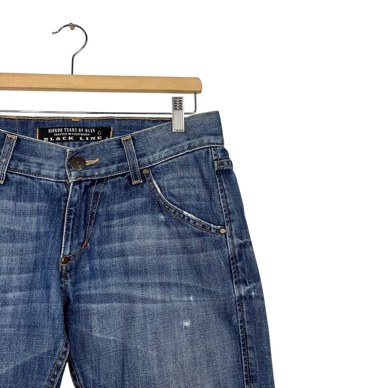 K&ouml;nnte beinhalten: Blaue Jeans aus Denim h&auml;ngen an einem Holzb&uuml;gel. Die Jeans haben eine verwaschene Optik, ein klassisches F&uuml;nf-Taschen-Design und einen Knopf- und Rei&szlig;verschluss. Das Etikett lautet "BLACK LINE".