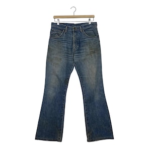 K&ouml;nnte beinhalten: Vintage blaue Jeans aus Denim mit ausgestelltem Bein-Design. Die Jeans h&auml;ngen an einem Holzb&uuml;gel vor einem wei&szlig;en Hintergrund. Der Denim hat ein verwaschenes Aussehen mit einigen Verf&auml;rbungen, was auf Abnutzung und Alter hindeutet. Die Jeans haben ein klassisches F&uuml;nf-Taschen-Design.