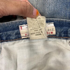 K&ouml;nnte beinhalten: Nahaufnahme eines blauen Jeans-Etiketts mit japanischem Text. Das Etikett enth&auml;lt Details wie den Produktcode M384 0102 N und HKD26. Die Jeans werden in China hergestellt.