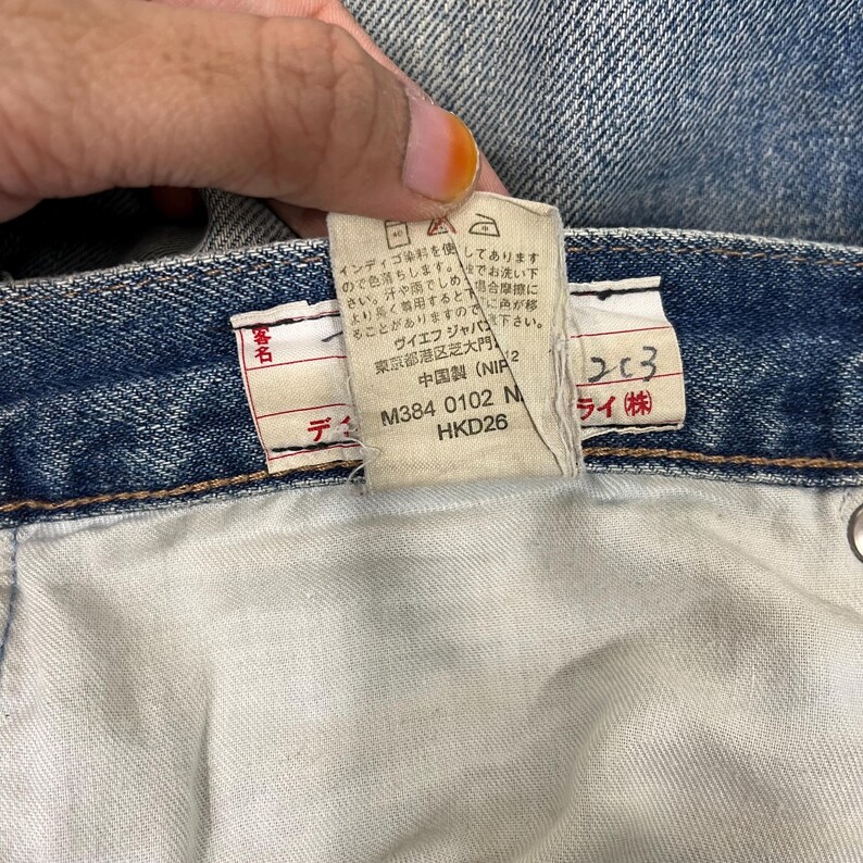 K&ouml;nnte beinhalten: Nahaufnahme eines blauen Jeans-Etiketts mit japanischem Text. Das Etikett enth&auml;lt Details wie den Produktcode M384 0102 N und HKD26. Die Jeans werden in China hergestellt.