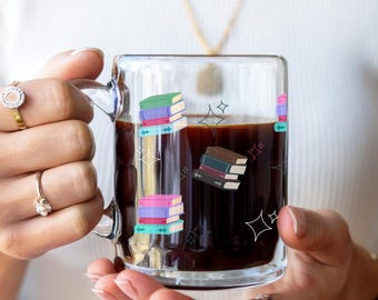 May include: A clear glass mug filled with dark liquid, possibly coffee or tea. The mug is decorated with colorful stacked book illustrations and starburst designs. The mug is held by a person.