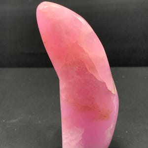 以下が含まれることがあります： 滑らかで湾曲した形状の、研磨されたピンク色の宝石。石はさまざまなピンクの色合いを持ち、明るい部分といくつかの暗いインクルージョンがあります。小さな金属製の留め金が底に付いています。宝石はおそらく装飾目的で使用されます。