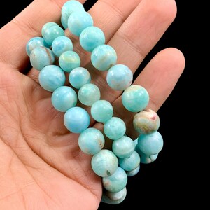 K&ouml;nnte beinhalten: Ein Armband aus hellblauen Edelsteinperlen. Die Perlen sind rund und variieren leicht in der Farbe, mit einigen, die Hinweise auf Gr&uuml;n und Braun aufweisen. Das Armband wird vor einem dunklen Hintergrund pr&auml;sentiert.