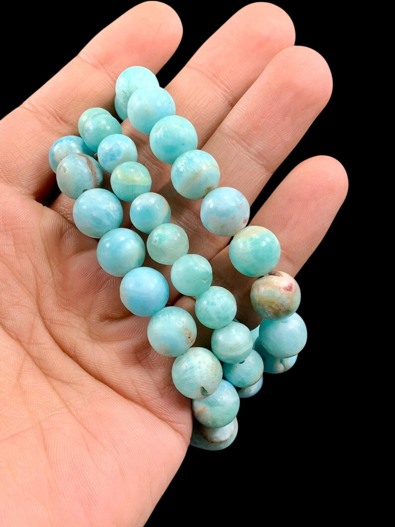 K&ouml;nnte beinhalten: Ein Armband aus hellblauen Edelsteinperlen. Die Perlen sind rund und variieren leicht in der Farbe, mit einigen, die Hinweise auf Gr&uuml;n und Braun aufweisen. Das Armband wird vor einem dunklen Hintergrund pr&auml;sentiert.