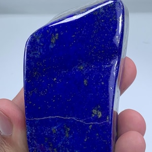 Op de afbeelding: Een gepolijste, diepblauwe lapis lazuli steen met een glad, glanzend oppervlak. De steen bevat natuurlijke insluitingen van goudkleurige pyrietspikkels verspreid over het oppervlak en een dunne, horizontale witte ader in het onderste gedeelte.