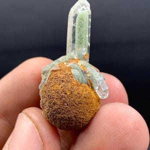 May include: A clear quartz crystal with a green interior, emerging from a rough, brown base. The crystal is held in a hand, showcasing its natural form and color variations. The base has a textured, earthy appearance.